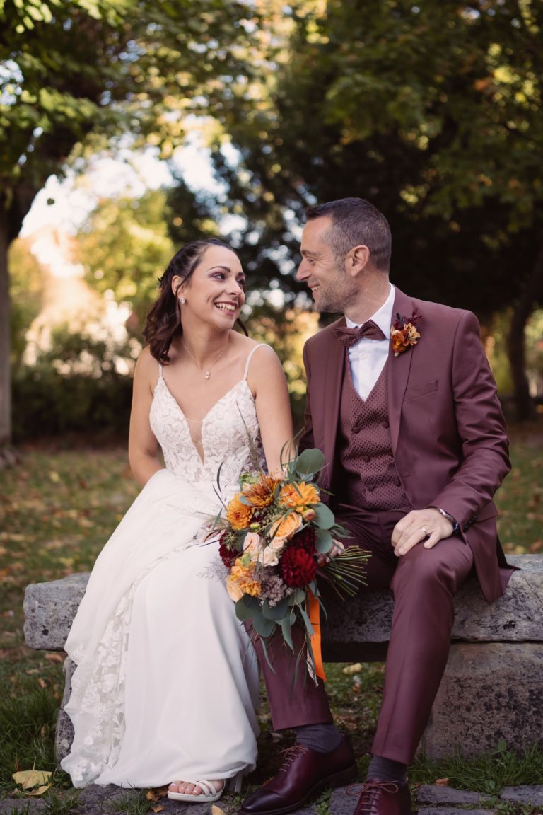 Photographie de mariage, deux mariés se regardent en souriant dans la forêt. La mariée tient son bouquet.