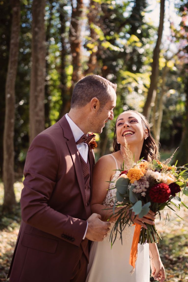 Photographie de mariage, deux mariés se regardent en souriant dans la forêt. La mariée tient son bouquet.